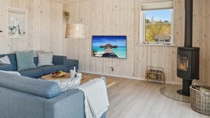 A cozy living room with a blue sofa, a television, a wooden wall, and a black stove. - "Engli" - 90m from the sea by Interhome (Væggerløse)
