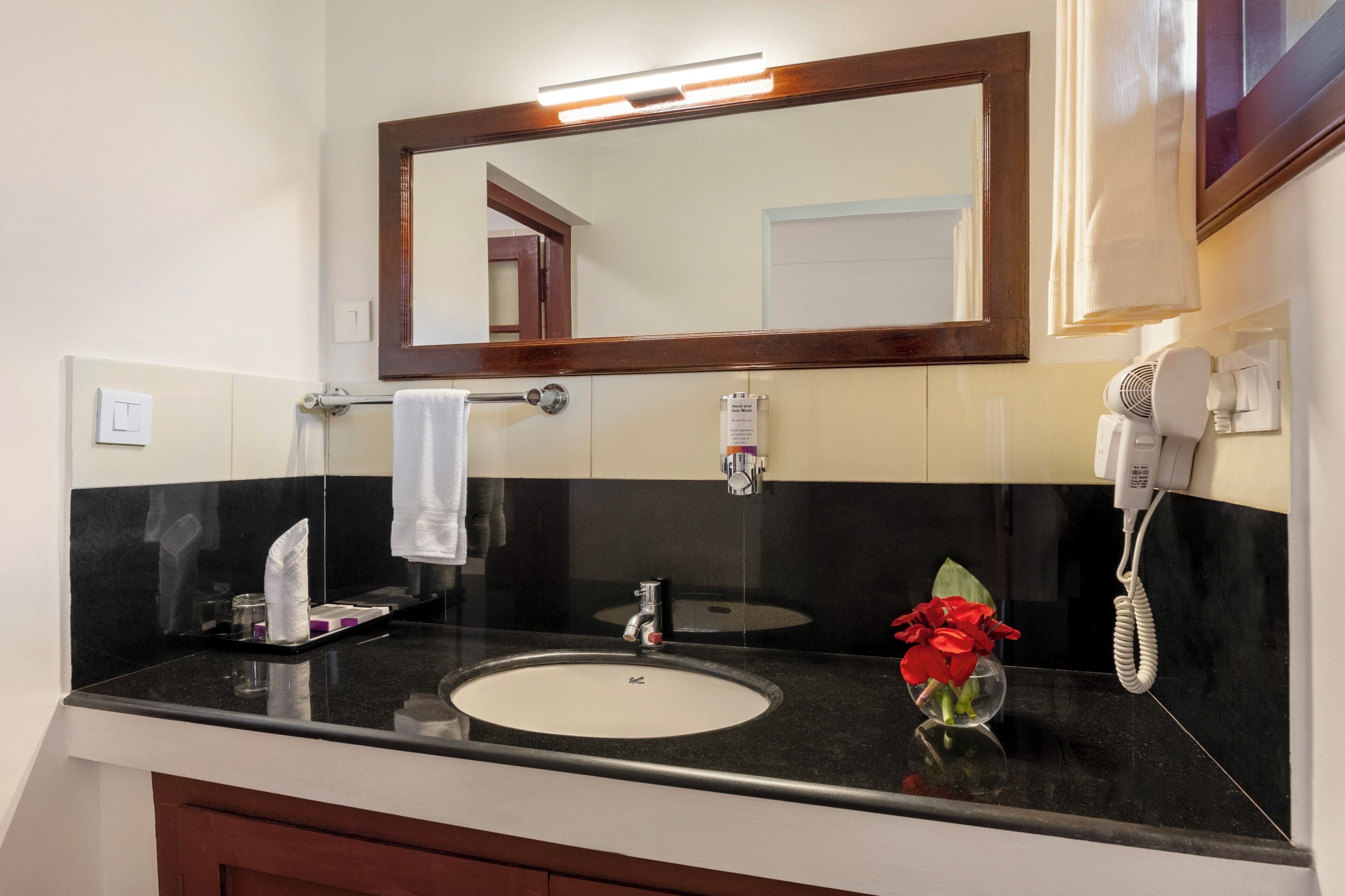 A bathroom with a black countertop, a white sink, a mirror, a hairdryer, and a vase with red flowers.