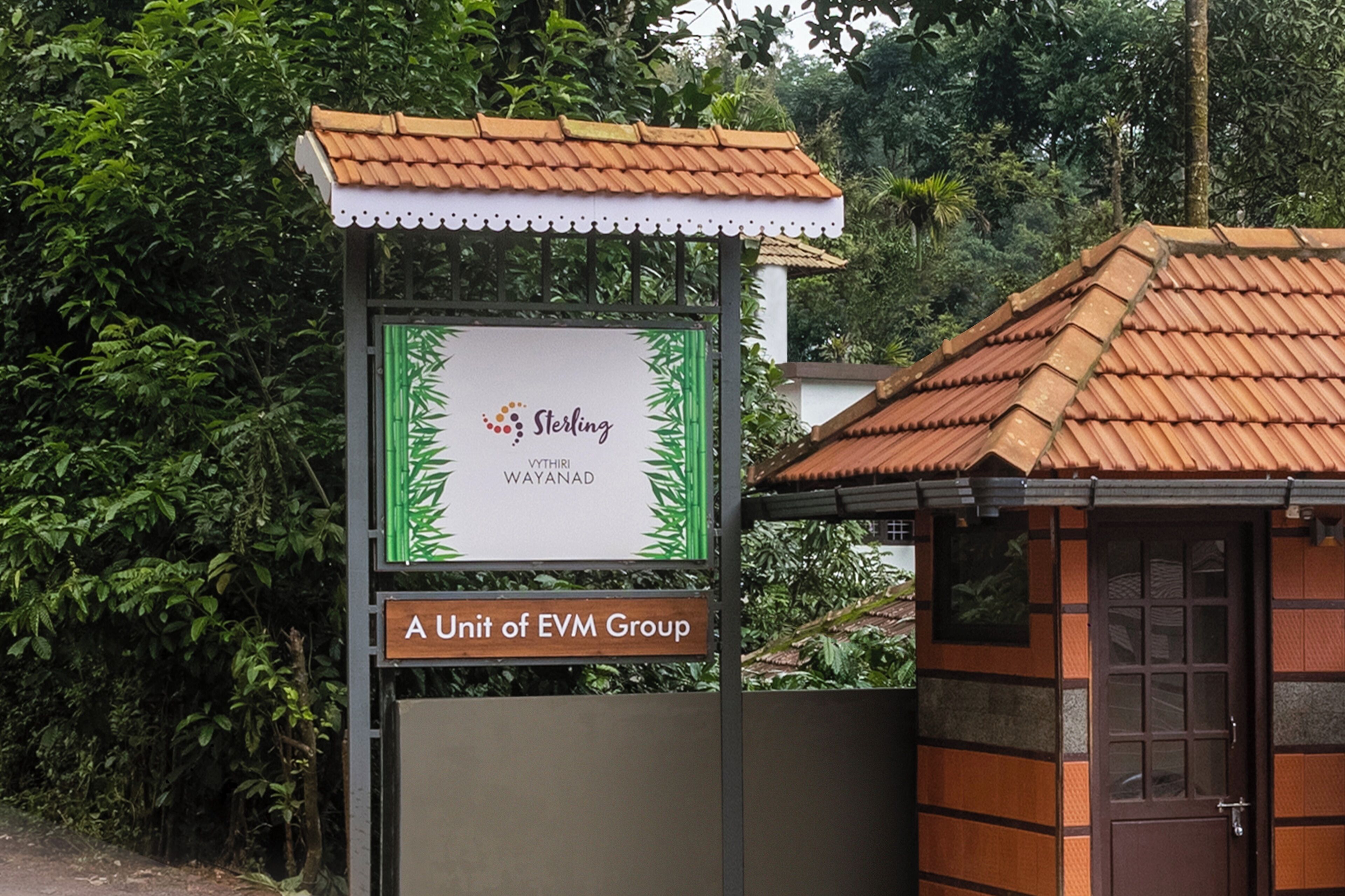 Señal de Sterling Vythiri Wayanad, una división de EVM Group, con un techo de tejas tradicionales y una pequeña estructura de madera cerca.