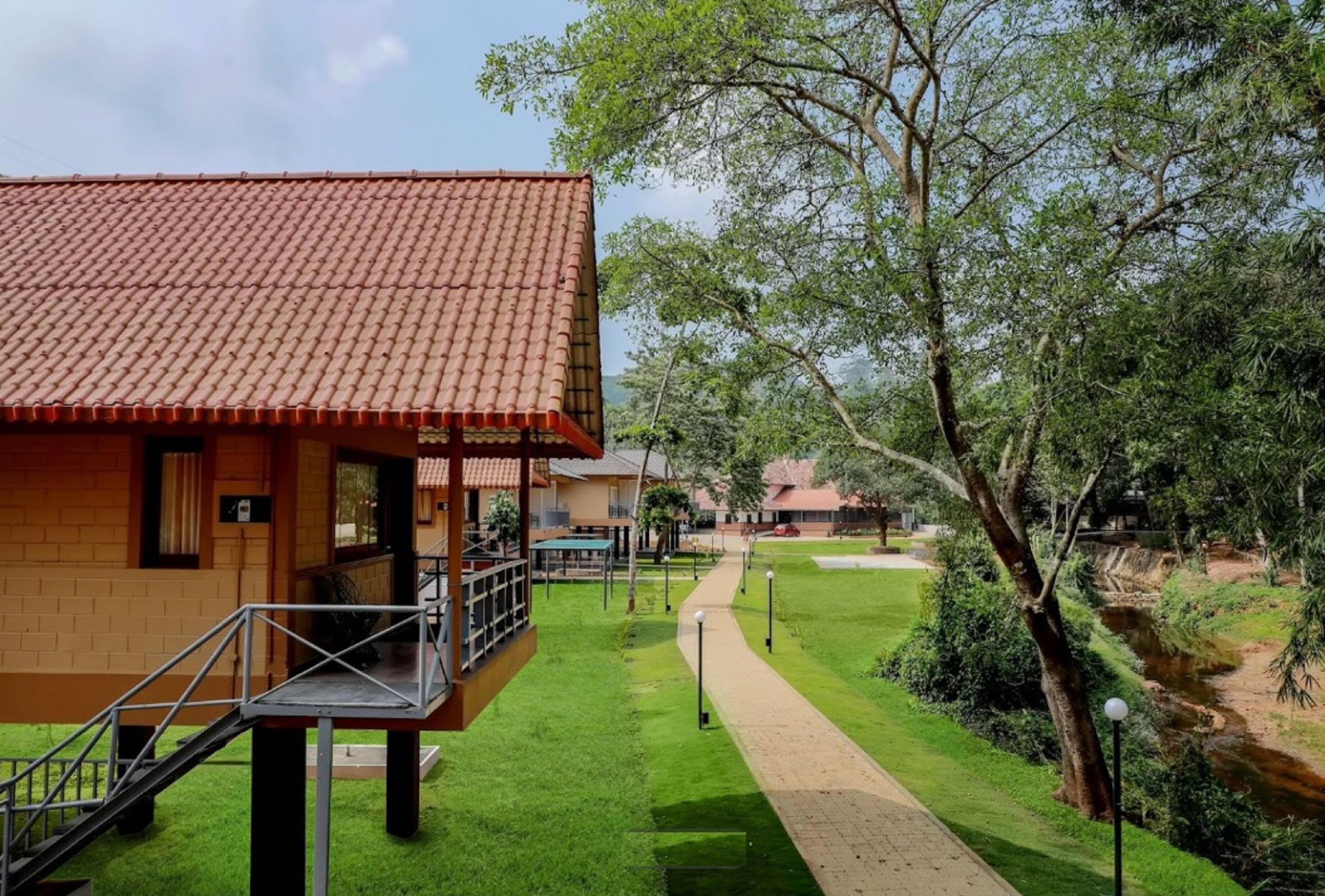 Een gebouw met een dak van rode tegels, een overdekt terras en een verhard pad dat leidt naar een gazon met bomen en een beekje.