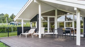 Ein überdachter Außenbereich mit Holzterrasse, weißen Liegestühlen und einem Tisch mit Stühlen.