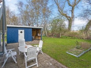 Eine blaue Schuppen mit Tür, ein kleiner Garten mit Tisch und Stühlen sowie ein Baum im Hintergrund.