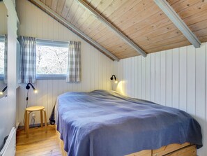 Ein Schlafzimmer mit Holzboden, einem Bett mit blauer Decke, einem kleinen Hocker und einem Fenster mit Vorhängen.