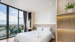 A modern hotel room with a large bed, a wooden floor, a view of the city, and a plant on a shelf. - Haya Bac Hai Apartments (Ho Chi Minh City)