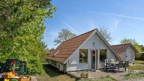 A small, single-story house with a sloped roof, a front porch with a table and chairs, and a small deck area.