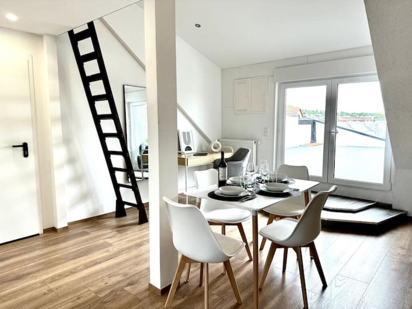 Un comedor con una mesa dispuesta para cuatro, una escalera en la esquina y vistas del exterior a través de un ventanal grande.