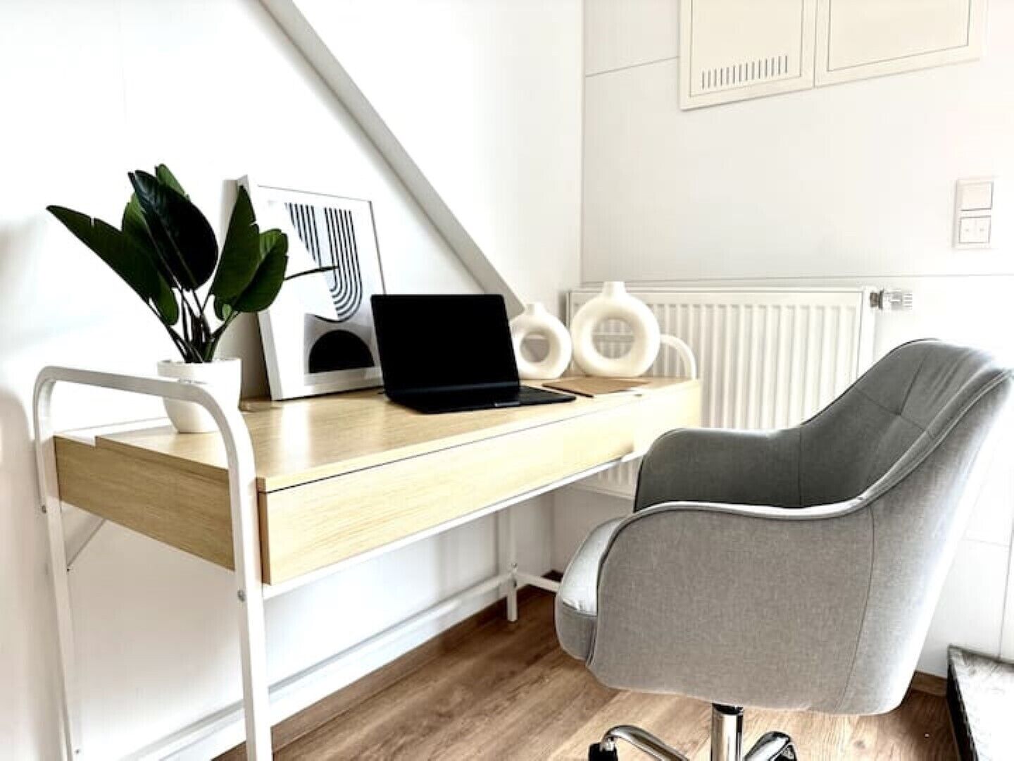 Un espace de travail moderne avec un bureau en bois, un ordinateur portable, une chaise de bureau grise, une plante en pot et un radiateur.
