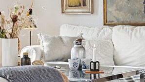 Ein modernes Wohnzimmer mit einem weißen Sofa, einem Glastisch und Dekorationselementen wie einer Kerze, einer Vase mit Trockenpflanzen und einem kleinen Hocker.