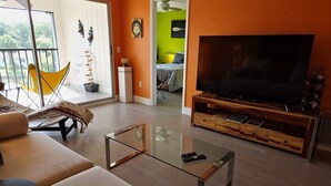 Ein Wohnzimmer mit einem Glastisch, einer Couch, einem gelben Sessel, einem Fernseher und einer Holz-Unterhaltungsanlage.