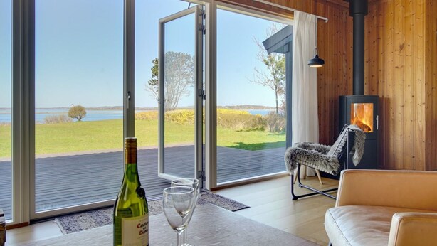 A room with a large sliding glass door leading to a deck with a view of a lake and trees. There is a fireplace with a log fire, a sofa, and a table with a bottle and glasses. - "Iadrik" - 85m to the fjord by Interhome (Skibby)
