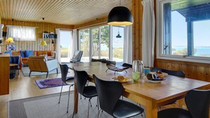 A cozy cabin interior with a wooden ceiling, a dining area with a table and chairs, a sofa, and a large window offering a view of the outdoors. - "Iadrik" - 85m to the fjord by Interhome (Skibby)