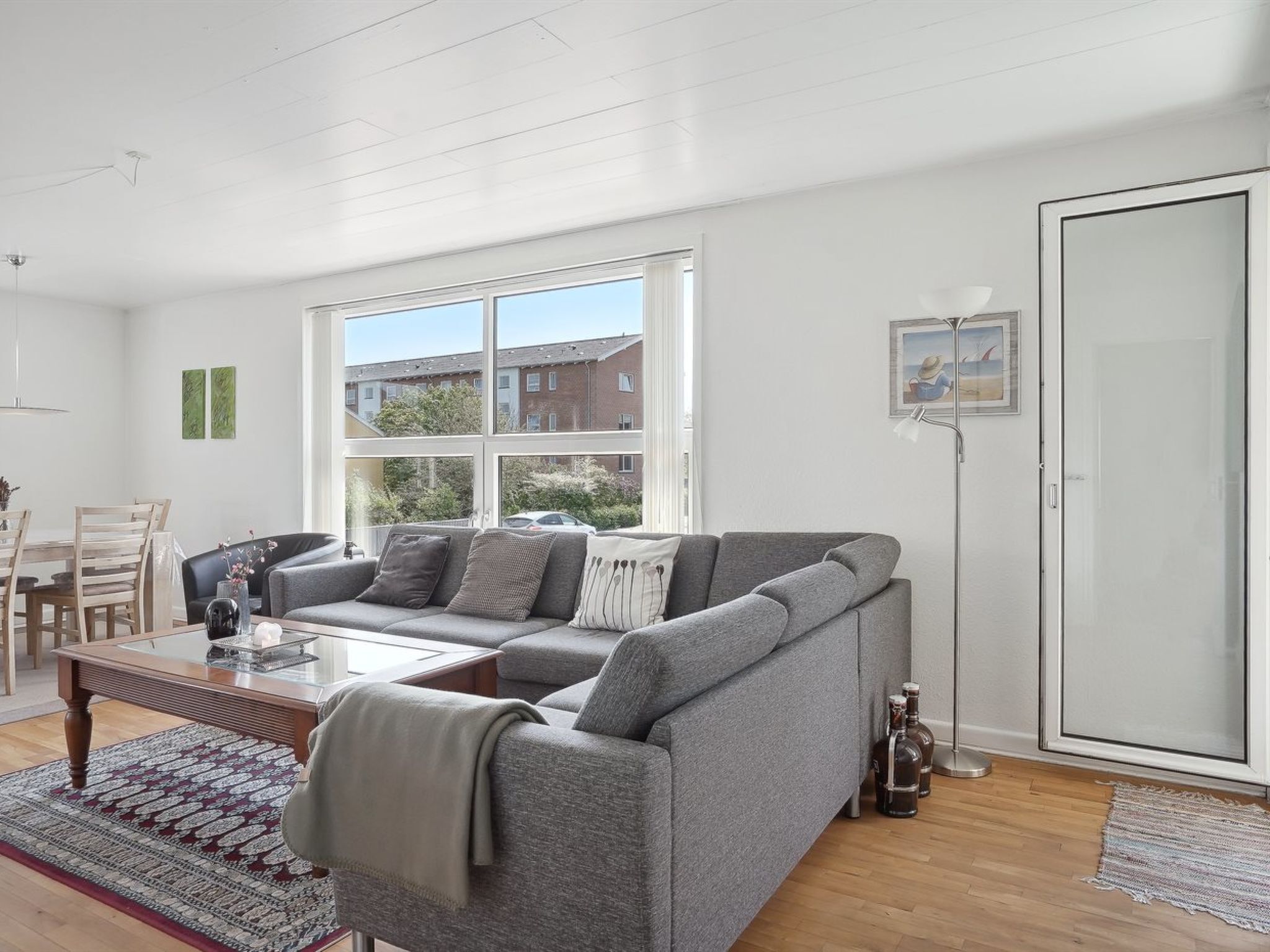 En moderne stue med en grå hjørnesofa, et træbord og et stort vindue med udsigt til bygninger og grønne områder.