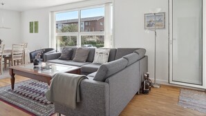 A modern living room with a grey sectional sofa, a wooden coffee table, and a large window offering a view of buildings and greenery.