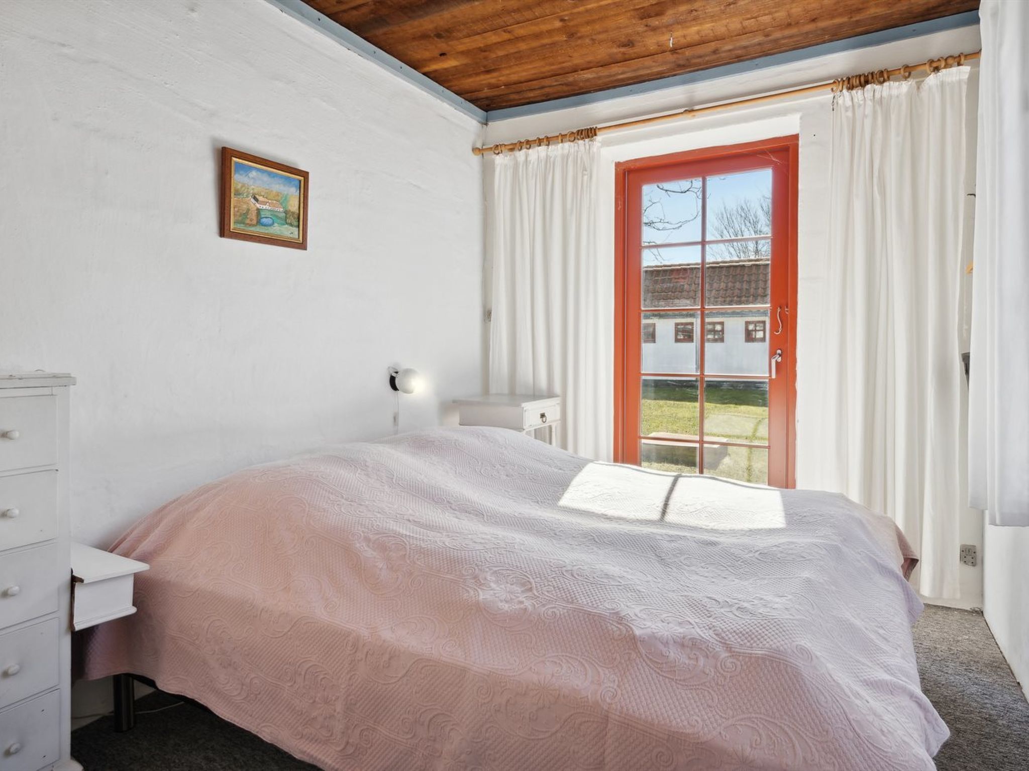 Ein Schlafzimmer mit Bett, Kommode, Nachttisch, Lampe, einem Fenster mit Vorhängen und einem Bild an der Wand.