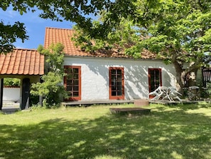 A white building with a red-tiled roof, two windows with red frames, and a small outdoor seating area with chairs and a table.