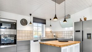 Una cocina moderna con armarios grises, una isla de madera, electrodomésticos de acero inoxidable, iluminación colgante y un ventanal que proporciona luz natural.
