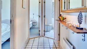 Een smalle gang met betegelde vloer die leidt naar een slaapkamer met een bed en nachtkastje.
