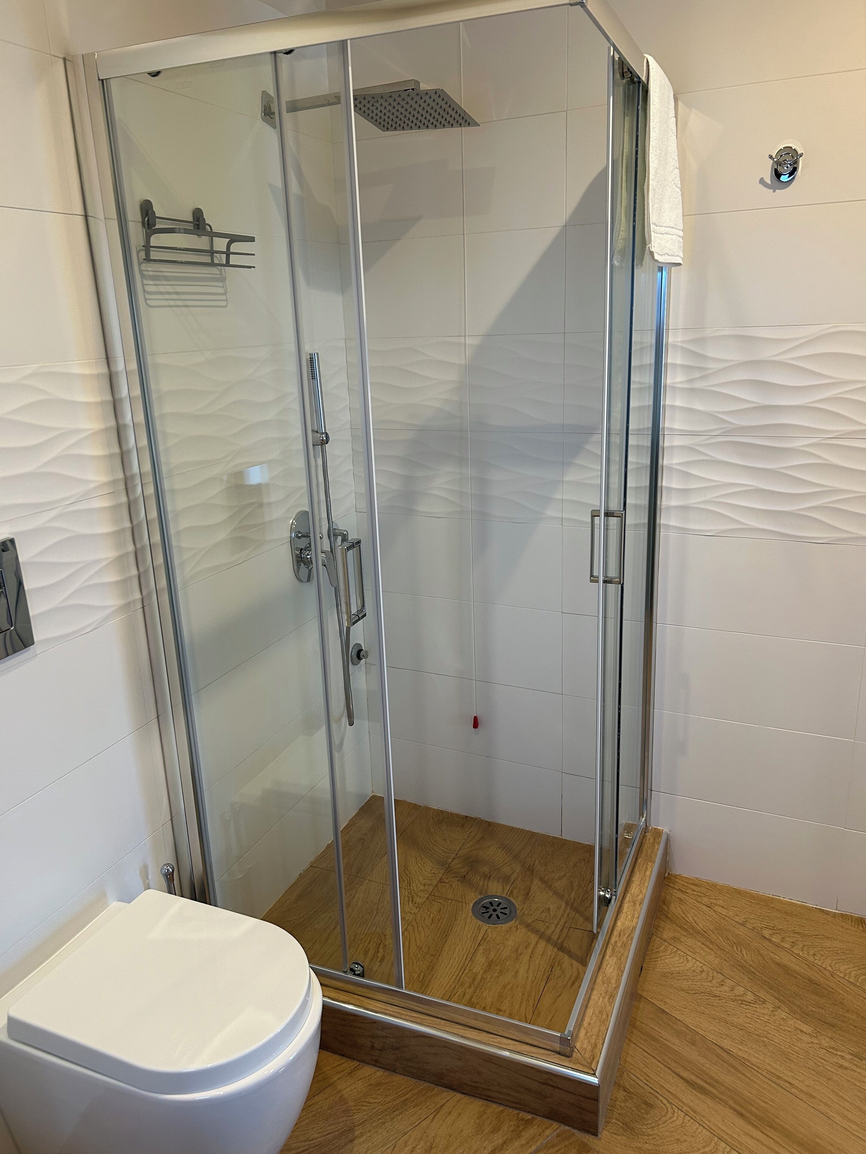 Une douche fermée par des parois de verre, avec un mur recouvert de carreaux blancs, un plancher en bois et des toilettes blanches.