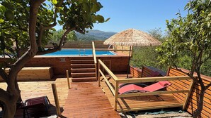 Une terrasse en bois mène à un espace piscine agrémenté d’un parasol en chaume, le tout entouré de verdure et d’arbres.
