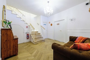 Un hall d’hôtel avec un escalier en bois, un extincteur, un lustre et un canapé marron recouvert d’une couverture colorée.