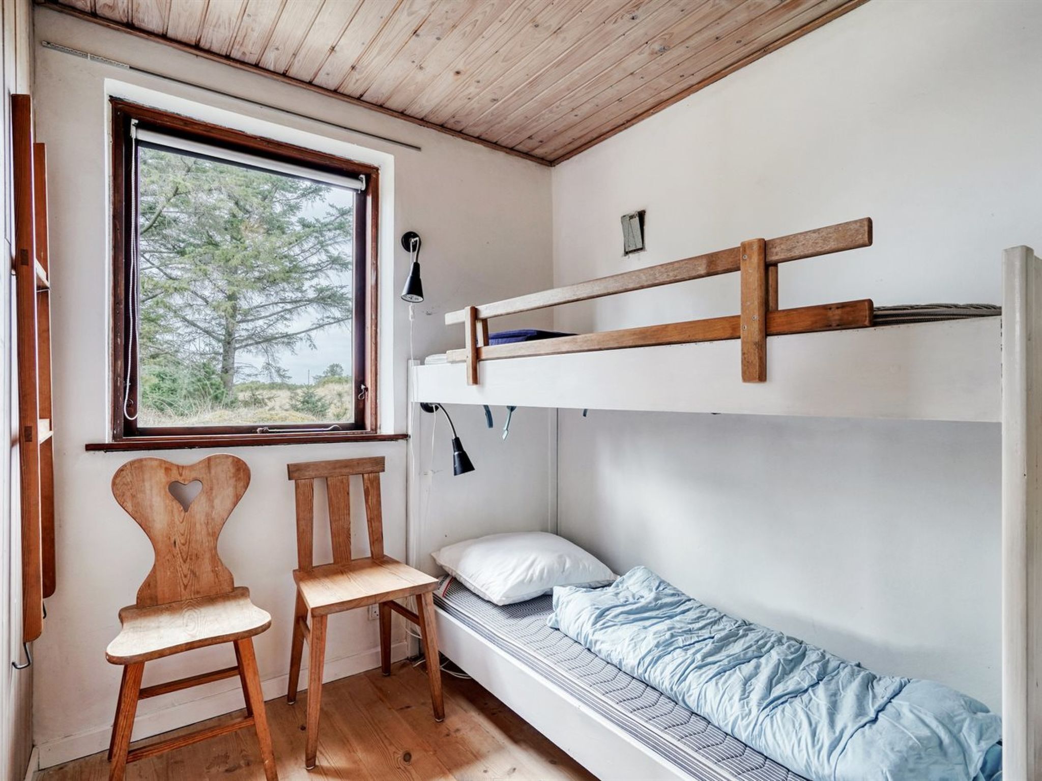 A bunk bed room with a window, two wooden chairs, and a bedside table.