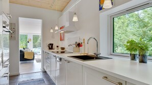 Una cuina moderna amb armaris blancs, un rentapeus negre, llums penjants i una finestra amb vista a vegetació.