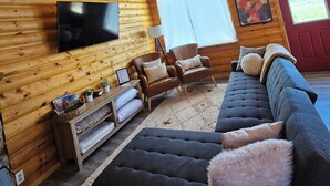 Interior de una cabaña de madera con un ciervo montado en la pared, un televisor de pantalla plana grande, un sofá y una ventana con vistas al exterior.