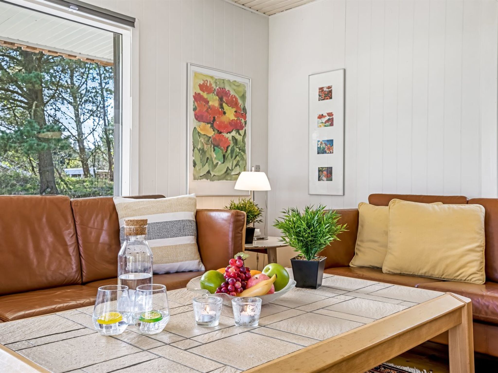 Un salon avec un canapé en cuir marron, une table basse avec des verres et des fruits, une lampe et un tableau accroché au mur.