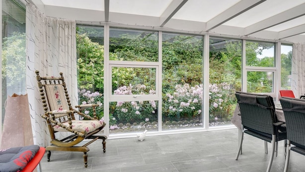 Une véranda dotée d’un toit en verre, une chaise en bois avec un coussin, une petite table et une vue sur une végétation luxuriante et des fleurs roses.