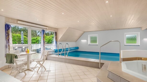 A modern indoor swimming pool with a sunlit patio area, a separate hot tub, and a view of greenery outside.