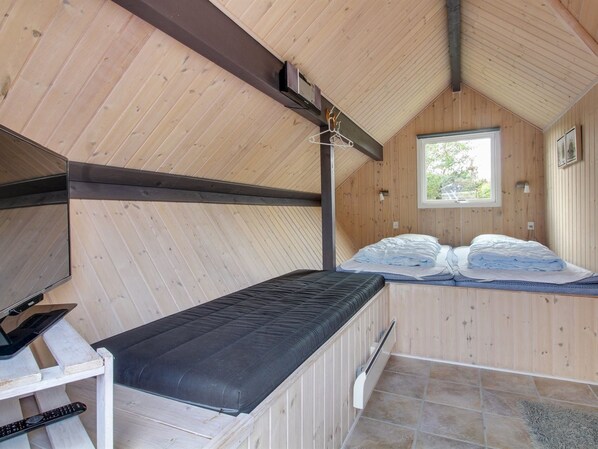 Ein kleiner, gemütlicher Raum mit Holzwänden und schrägen Decken. Es gibt ein Bett mit schwarzer Matratze, einen kleinen Tisch mit einem Laptop und einer Fernbedienung sowie ein Fenster, das natürliches Licht hereinlässt.