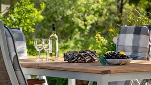 Une table en bois, installée à l’extérieur, est dressée avec une bouteille de vin blanc, deux verres, un bol d’olives et une bûche décorative servant de centre de table.