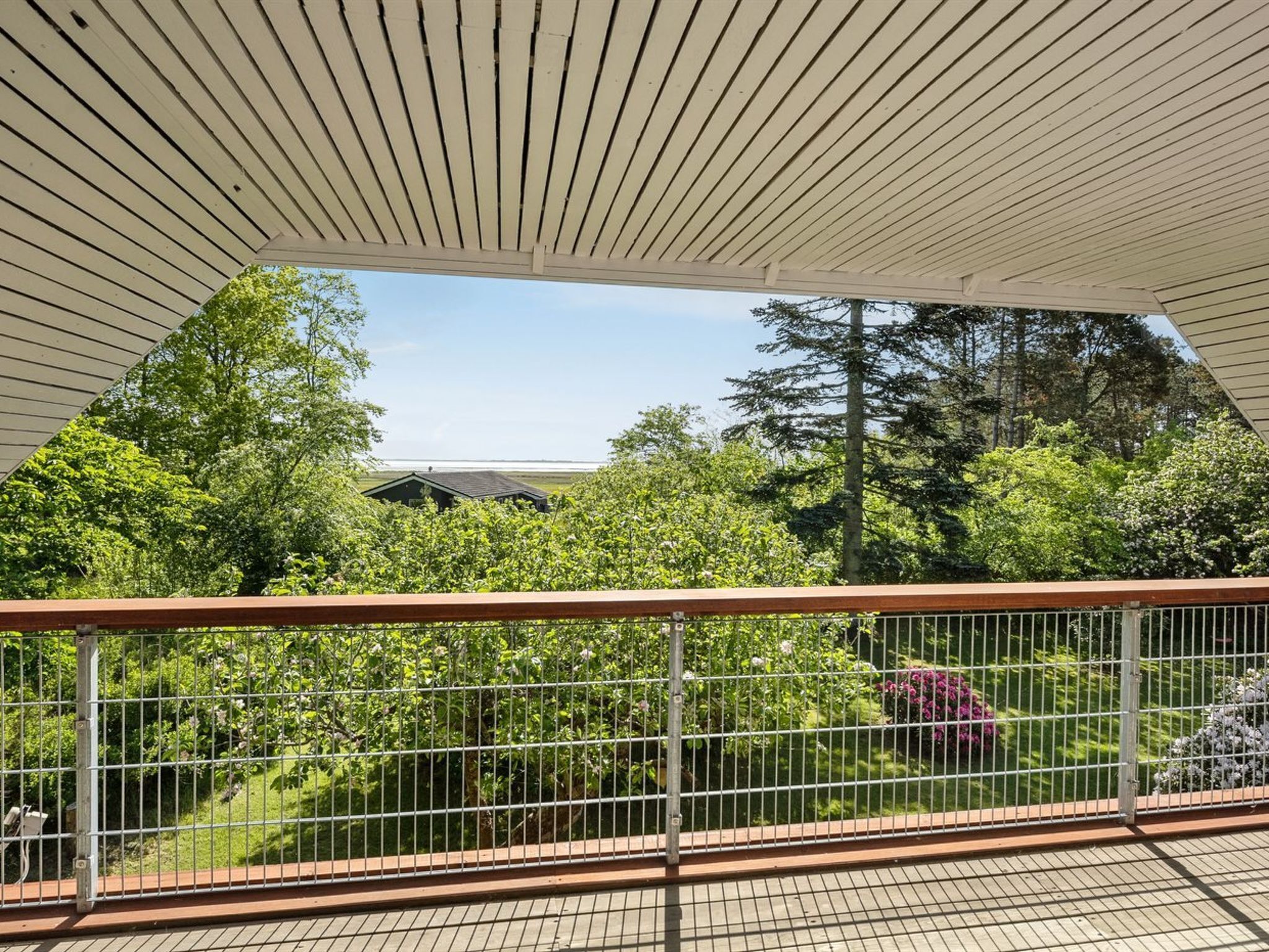 Un espace extérieur abrité doté d’une terrasse en bois, d’une rambarde en métal et offrant une vue sur un paysage verdoyant, avec des arbres et un bâtiment visible au loin.