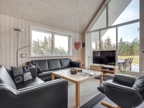 Un salon moderne avec un canapé, une table basse et un téléviseur, de grandes fenêtres et une vue sur l’extérieur.