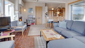 Un grand espace de vie comprenant un canapé, un téléviseur et une table à manger. Le sol est en bois et une grande fenêtre laisse entrer la lumière naturelle.