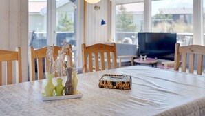 Un coin repas avec une table dressée pour deux personnes, agrémenté d’un vase contenant des fleurs séchées, d’un plateau avec un panier tressé et d’un téléviseur en arrière-plan.