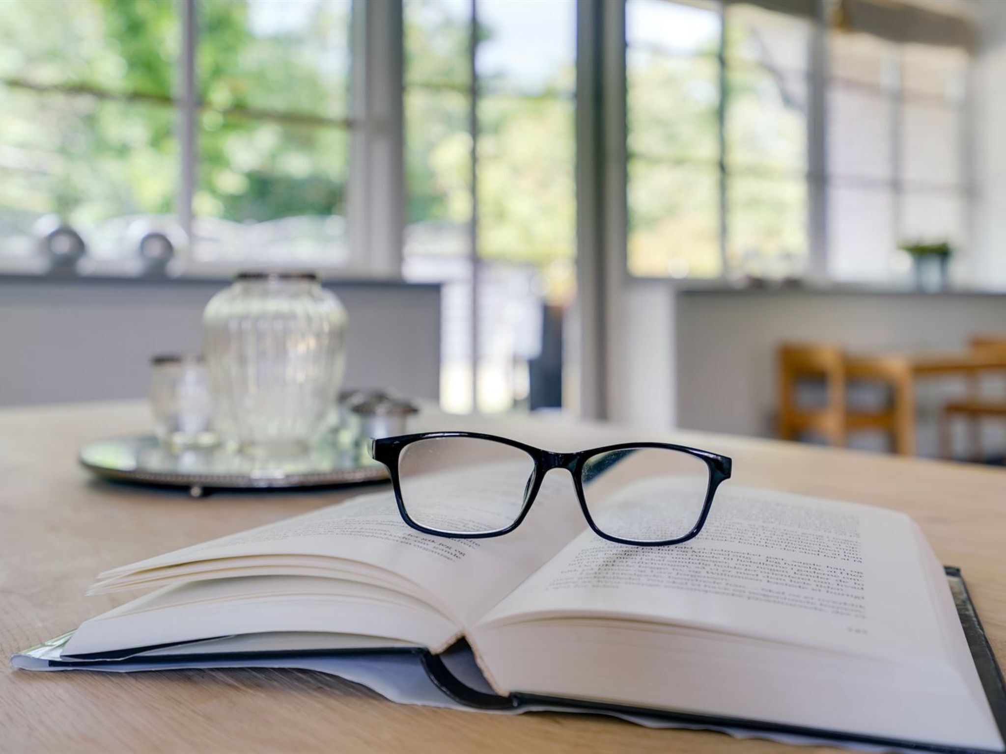 An open book on a wooden table with a pair of glasses on top, a glass container, and a window with a view of greenery.