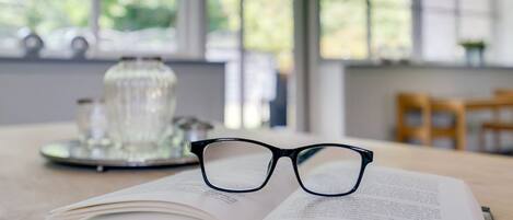 An open book on a wooden table with a pair of glasses on top, a glass container, and a window with a view of greenery.