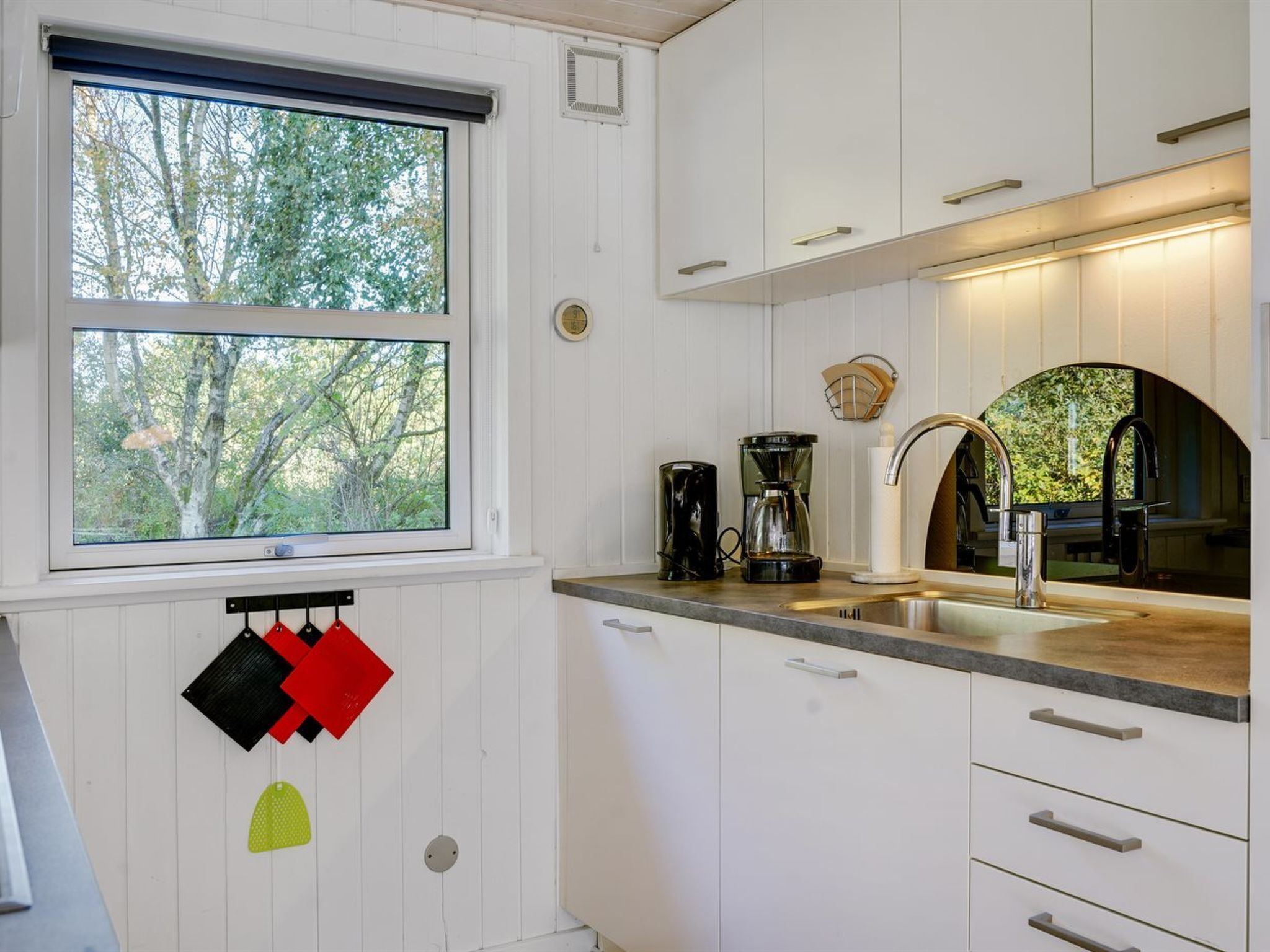 Een moderne keuken met witte kastjes, een groot raam met uitzicht op bomen, een koffiezetapparaat en een spoelbak met een moderne kraan.
