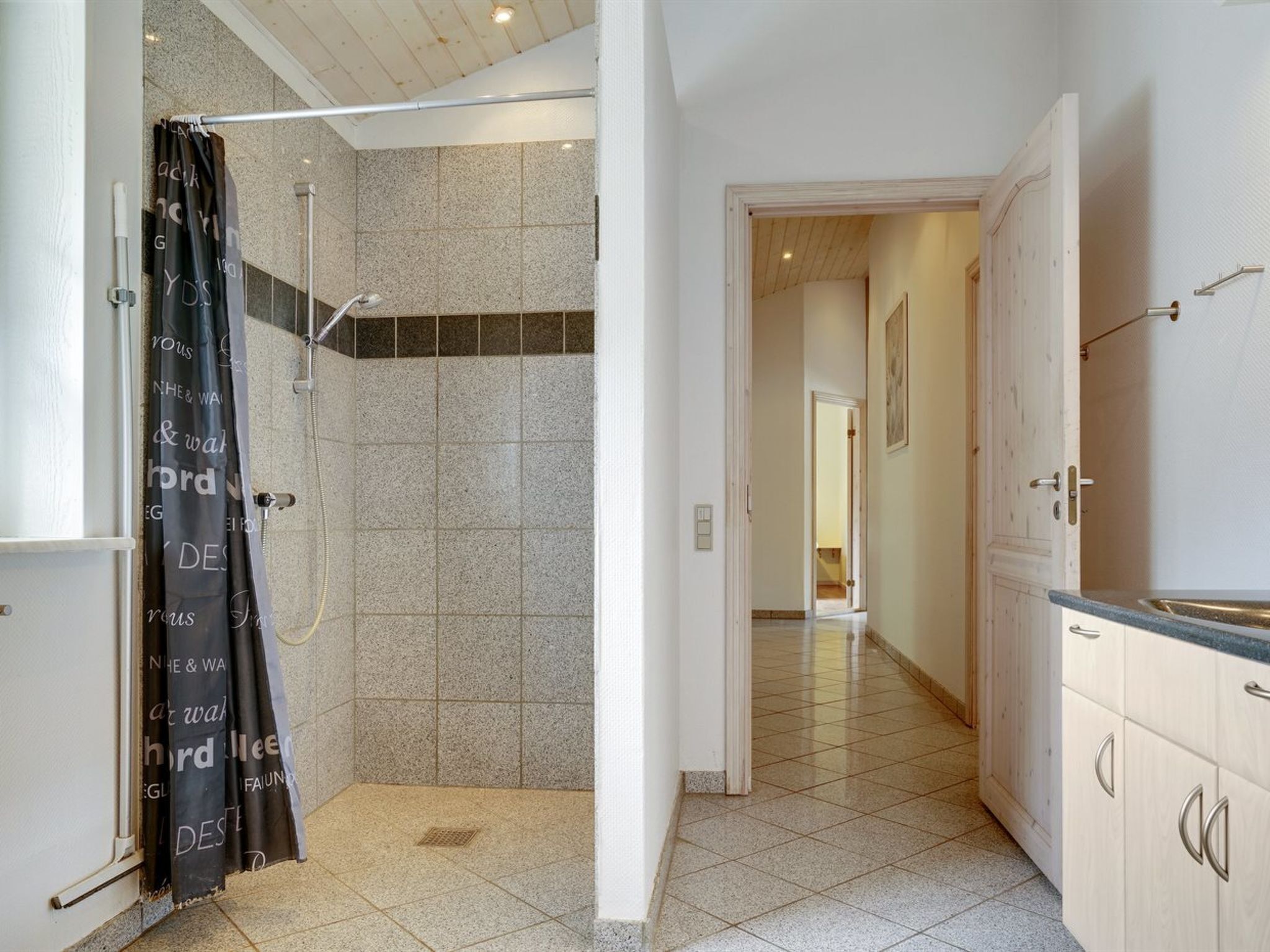 Une salle de bain avec douche, des murs carrelés et un couloir menant à une cuisine.