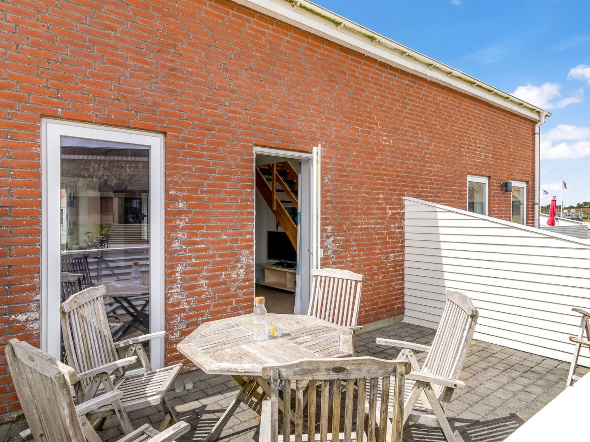 En murstensbygning med en patio, der har et træbord og stole, en trappe og en skydedør i glas.
