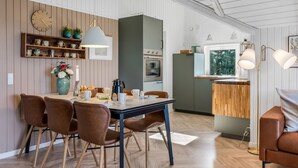 Moderni keittiö, jossa on puiset kaapit, ruokailutila pöytineen ja tuoleineen sekä ikkuna, josta avautuu näkymä vehreään maisemaan.