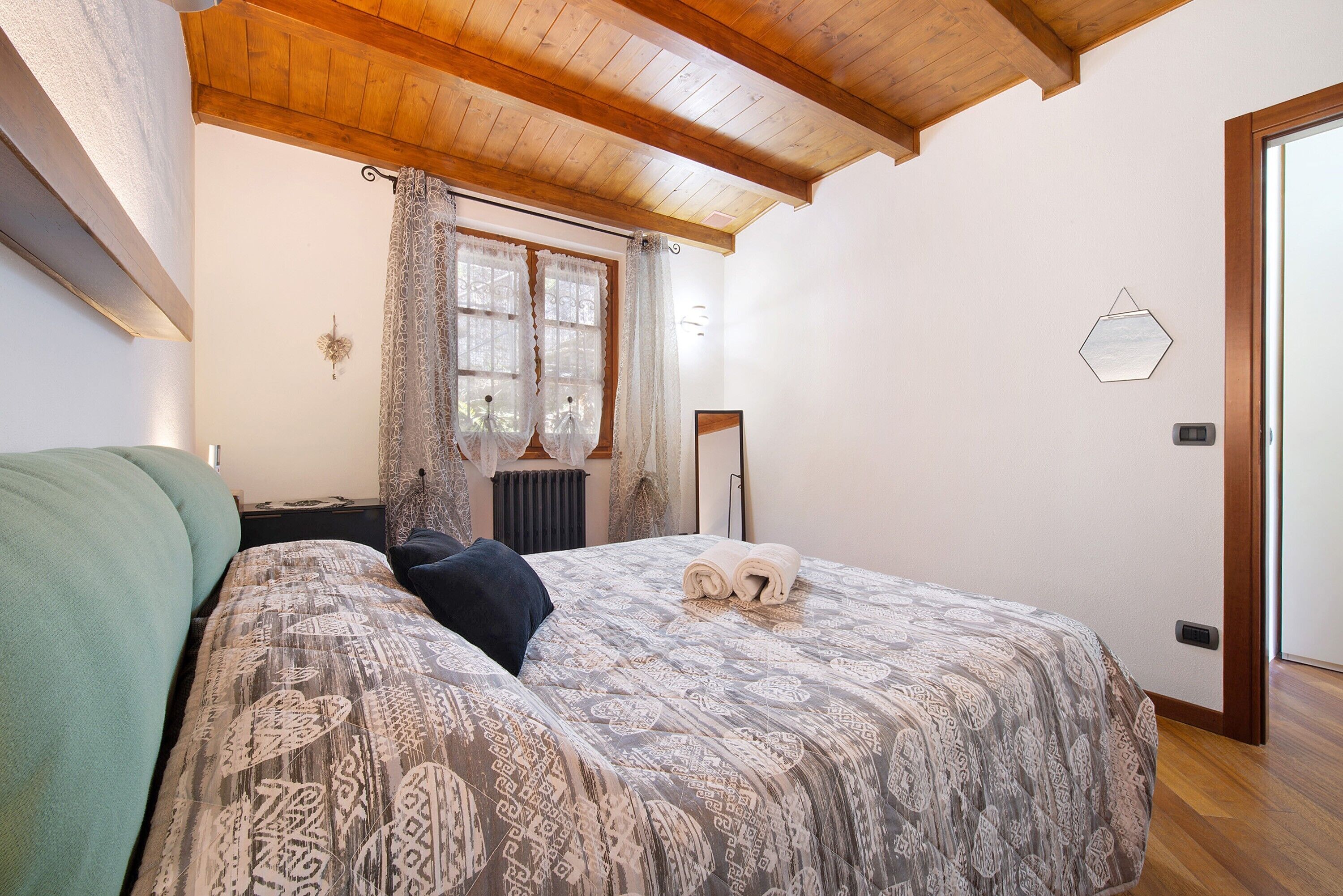 A bedroom with a bed, a window with curtains, a radiator, and a wooden ceiling.