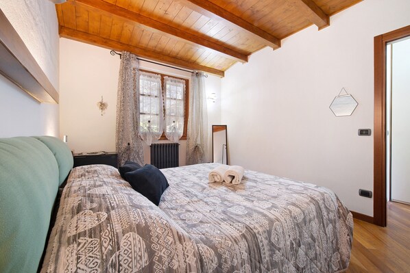 Ein Schlafzimmer mit einem Bett, einem Fenster mit Vorhängen, einer Heizung und einer Holzbalkendecke.