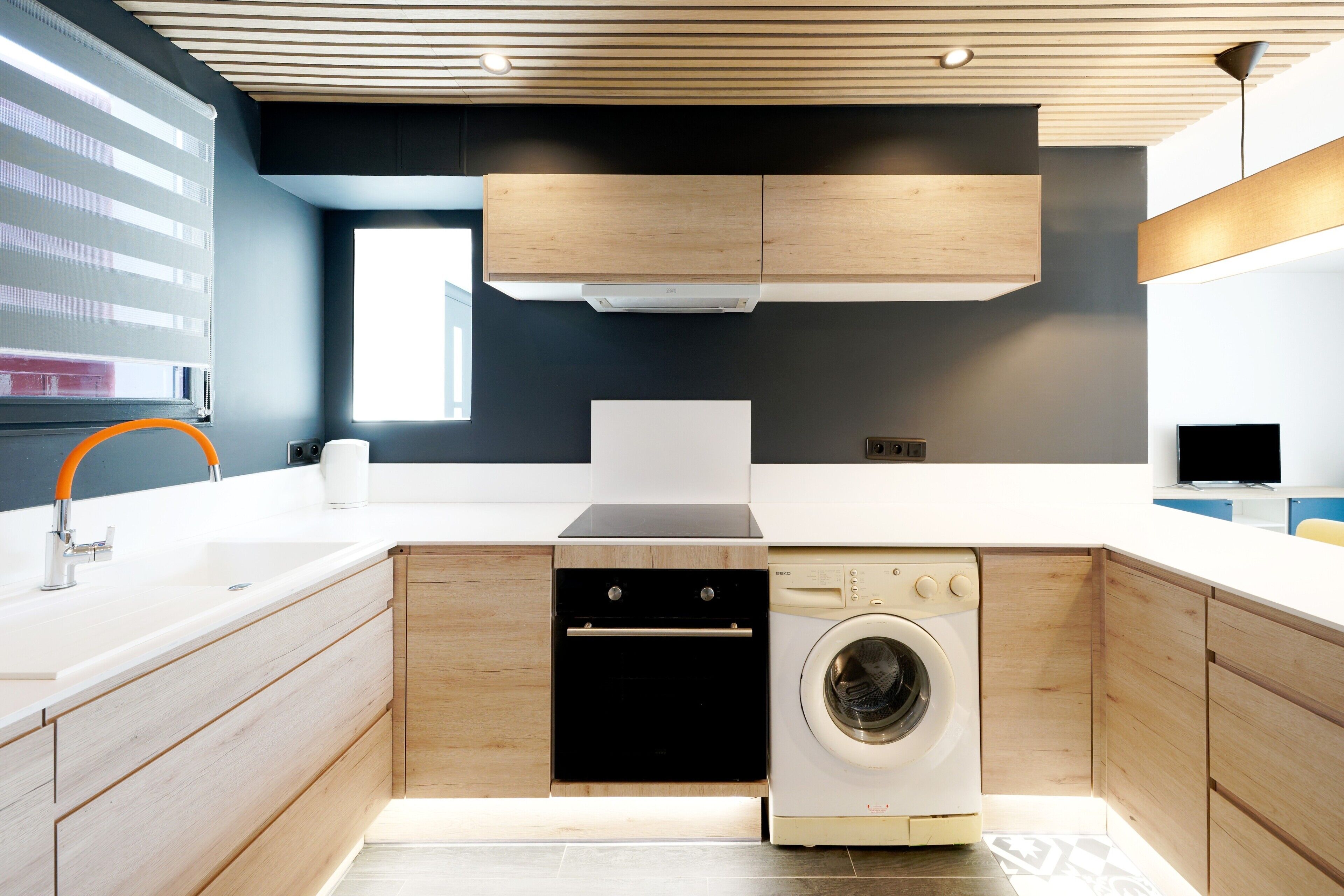 A modern kitchen with wooden cabinets, a built-in oven, a washing machine, and a large window.