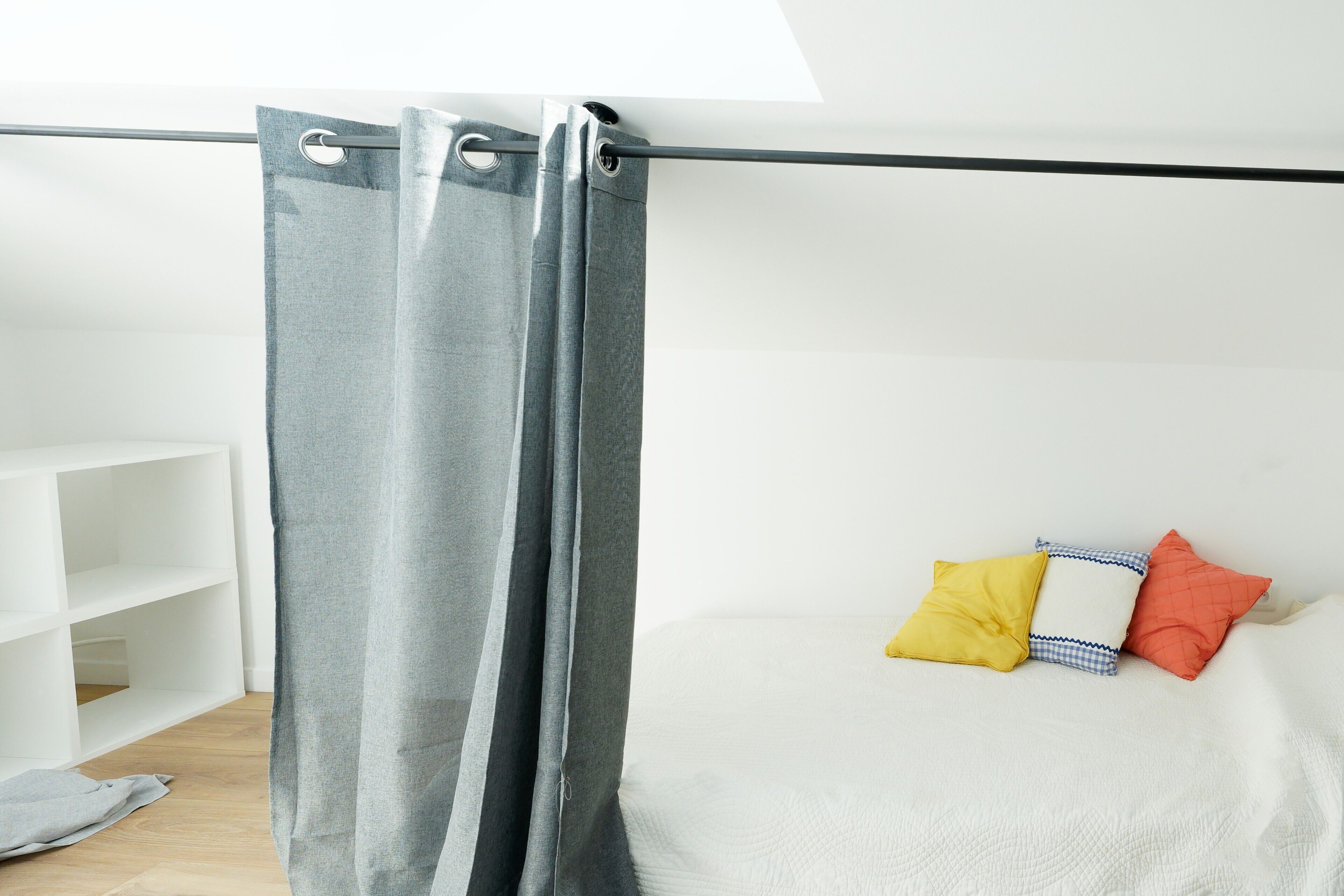 Una habitación con cortina gris, cama blanca con almohadas, estantería blanca y tragaluz.