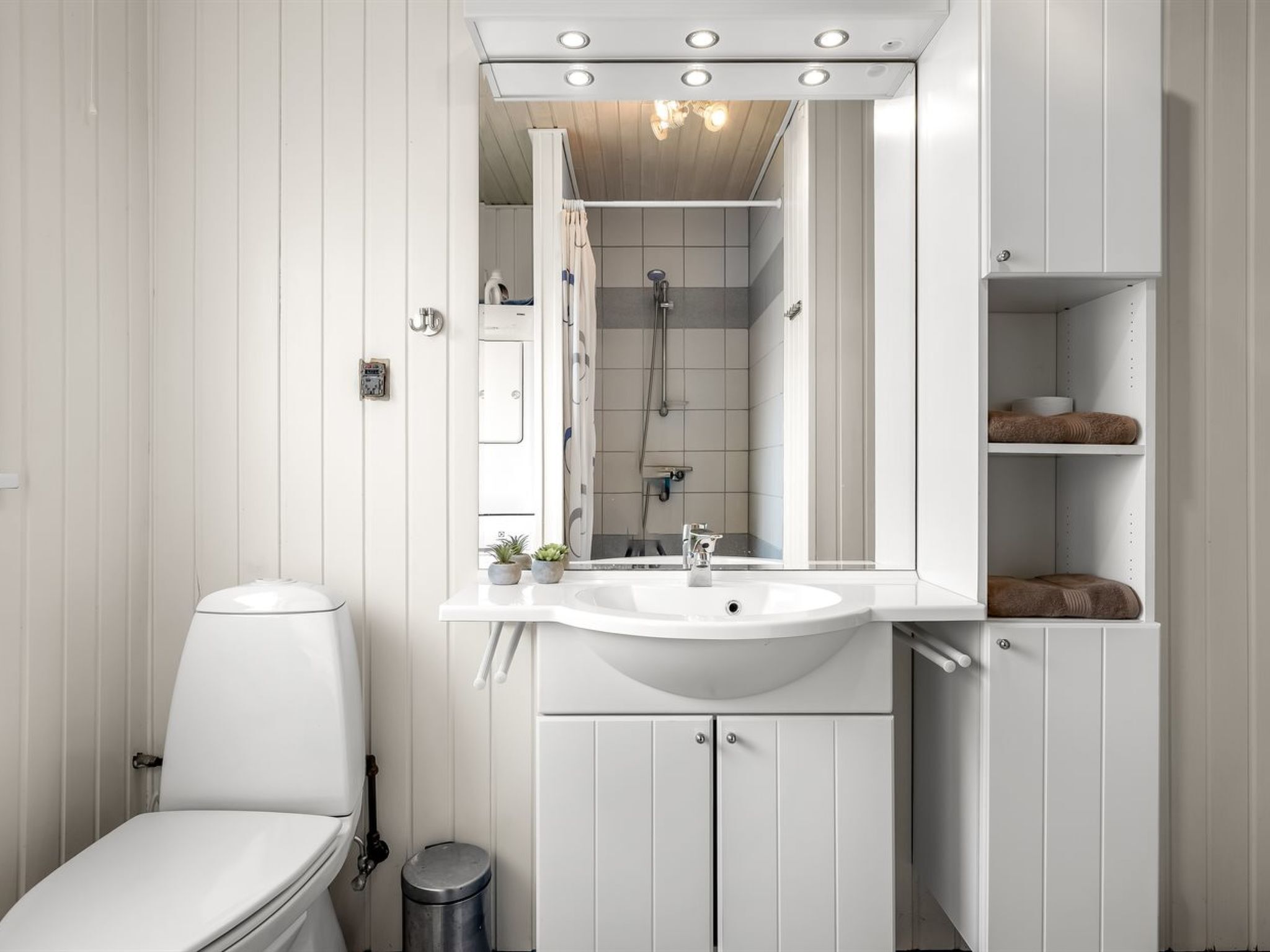 A bathroom with a toilet, sink, mirror, shower, and storage cabinets.
