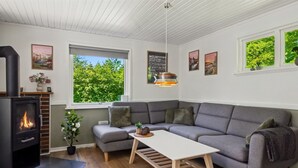 A modern living room with a built-in wood stove, a grey sectional sofa, a wooden coffee table, and large windows offering a view of greenery.