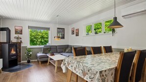 A modern living room with a fireplace, a sofa, a dining table with chairs, and a large window with a view of greenery.