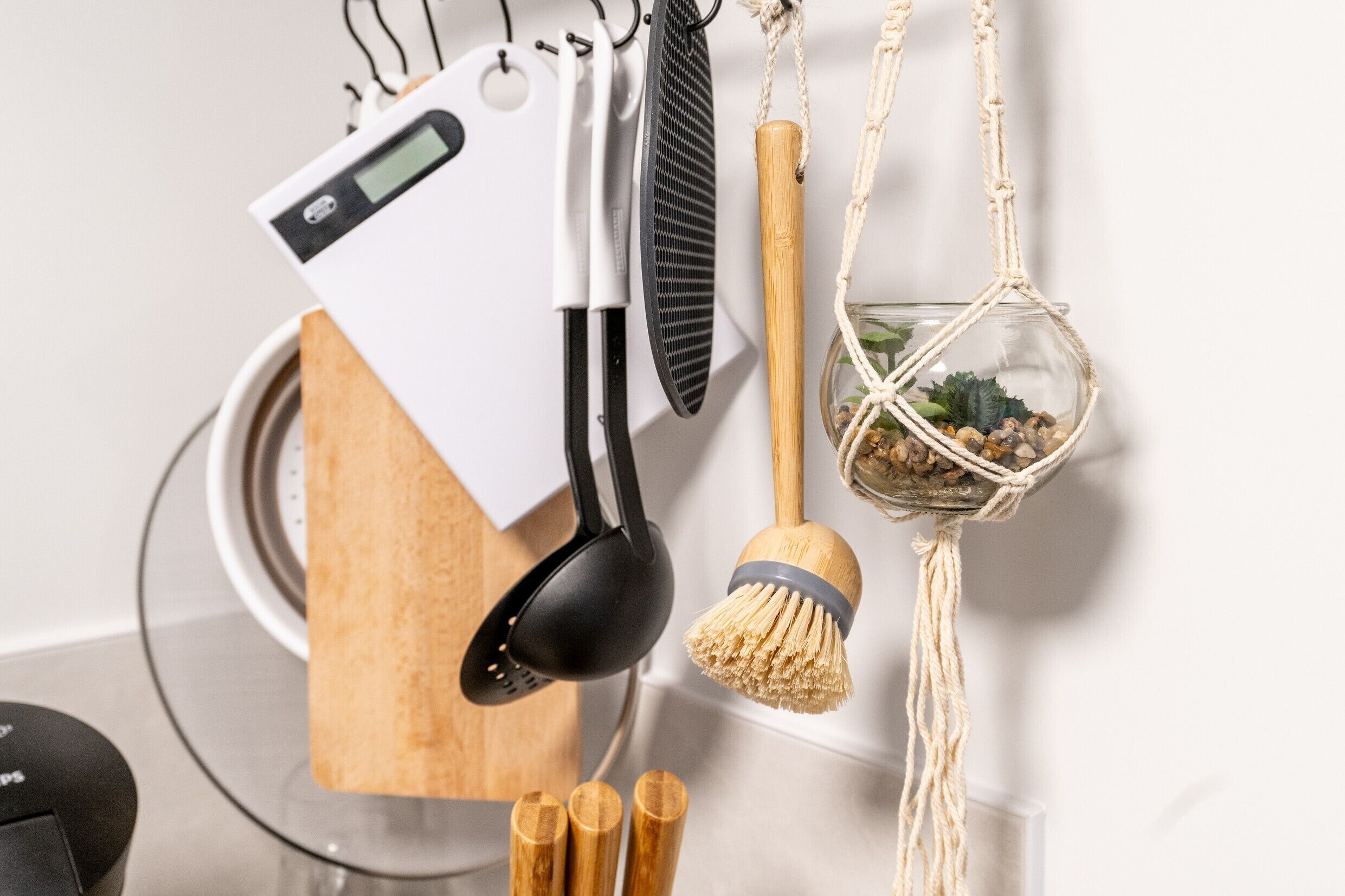 Des ustensiles de cuisine sont accrochés au mur, notamment une balance numérique, une spatule, un fouet et un pot suspendu en macramé dans lequel est plantée une petite plante.